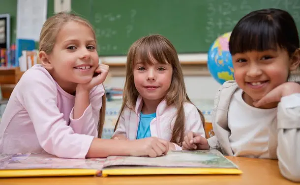 Happy schoolgirls reading a fairy tale to their classmate while looking at the camera