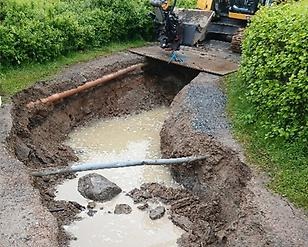 Grävarbete vid vattenfylld grop där rör blottas och maskin arbetar intill buskar på grusväg med lera.
