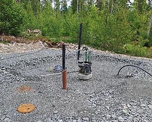 Grusplan med två rör och pump, slang på marken, skog i bakgrunden med kablar och grå sten yta syns.