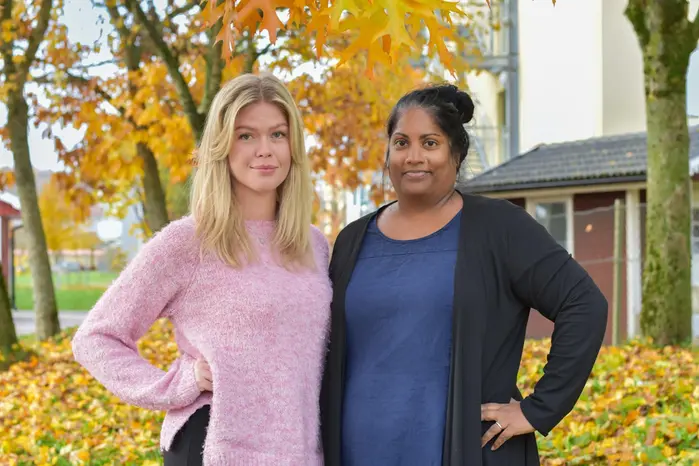 Två personer står framför träd med färgglada höstlöv och en byggnad i bakgrunden.