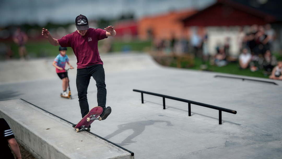 Person gör ett skateboardtrick vid kanten av en betongramp i en skatepark; en annan åker kickbike i bakgrunden.